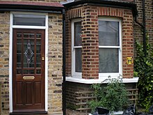 A brown brick building. Visible is a door at left, with a bowfront window at right.
