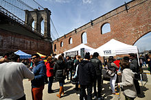 People crowd around white tents in the foreground next to a red brick wall with arched windows. Above and to the left is a towering stone bridge.