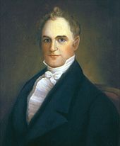 A man with receding, red, curly hair wearing a white shirt and tie and black jacket