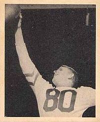Black and white photograph of Armstong in a light-colored number 80 jersey reaching up with his right hand to catch a football