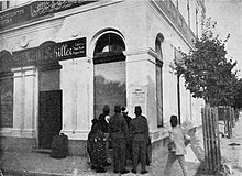 Photo of large white building with one signs saying "Moritz Schiller" and another in Arabic; in front is a cluster of people looking at poster on the wall.