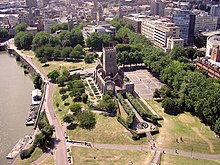 The walls and tower of an old ruined church set in a paved area and surrounded by a park. On the left is water with some pontoons moored and in the background office blocks, streets and church spires.