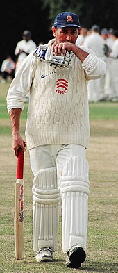 A cricket player facing the camera, walking off the pitch with his bat in his right hand, gloves in his left, wiping his mouth with his left hand