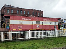 A railroad caboose parked on a siding behind a white picket fence. It reads "PROVIDENCE & WORCESTER" on its side.