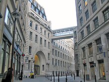 A narrow road with tall buildings of grey stone on both sides. The building on the left has a large entrance archway.