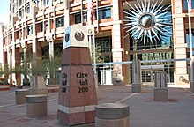 photos showing the short obelisk signage showing City Hall, and topped with the seal of the city, a stylized maroon phoenix. The semi-circular front of the building in the background, adorned with a stylized sunburst.