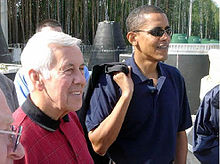 Gray-haired man and Obama stand, wearing casual polo shirts. Obama wears sunglasses and holds something slung over his right shoulder.