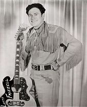 A dark-haired man wearing a neckerchief, a shirt with fringes dangling from the sleeves, and pants with a guitar pictured on them, smiling broadly while leaning on a guitar