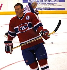 An ice hockey player in his forties stands on the ice wearing a red jersey with horizontal blue and white stripes and his stick held across his waist. He is concentrating on something off in the distance.