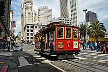 A red cable car operating on a city street, surrounding by tall buildings