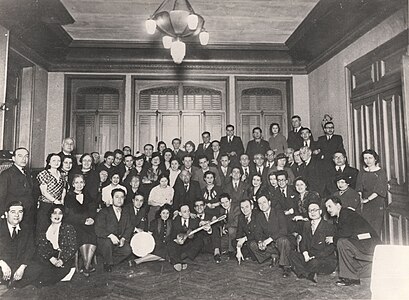 Photographie en noir et blanc de l'intérieur d'une pièce où pose sur plusieurs rangées une cinquantaine de personnes.