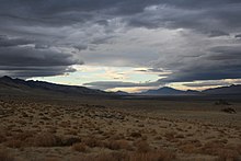 A landscape shot of a long, dry valley. The sky is partially clouded over but blue sky breaks through in patches. It is a showcase of Nevada's natural beauty.