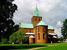 En forma de cruz, con techo de cobre, iglesia de ladrillo en estilo Románico con ventanas con arcos redondeados y un domo ábside. La larga torre tiene ventanas de estilo gótico puntiagudas.