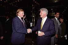 Donald Trump shakes hands with Bill Clinton in a lobby; Trump is speaking and Clinton is smiling, and both are wearing suits.
