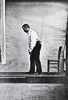 黑白照片中的帕金森氏症患者走路時身體佝僂，照片由他的左邊拍攝，而他的後方有一張椅子。