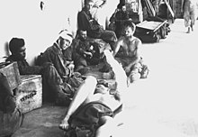 Several men are sitting and laying down near a building. Some are bandaged, and others have no shirts. Various boxes and cots are nearby.