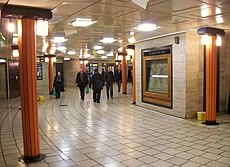 View of part of circular concourse with cream tiled floor, coffered flat ceiling and travertine stone walls. Faceted orange columns support bronze light fittings.