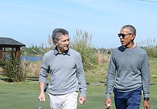 Obama in casual attire stands with a man in a golf course.