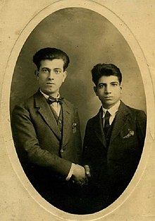 Photographie en noir et blanc de deux jeunes hommes en costume se serrant la main en regardant l'objectif.