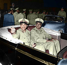 Two smiling men in military uniform seated in an open-top automobile. The first man on the left is pointing his hand in a gesture. Behind the automobile are men in uniform walking away from the vehicle