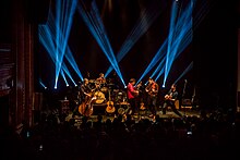 Five male musicians perform on stage in front of a standing audience, behind them a dozen lights project blue lines upward.