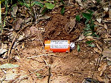A red palm-sized cylindrical metal container, lying in a shallow freshly dug hole in the ground, is labeled "ENREGISTREUR DE VOL / NE PAS OUVRIR".