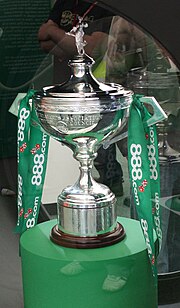 A silver cup-shaped trophy with a trumpet base, a Greek shepherdess finial on top, and two deco square-section handles at the sides with green sponsor ribbons tied to them; the trophy is sitting on a green cylindrical plinth