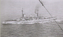 A light gray battleship steams in choppy seas.