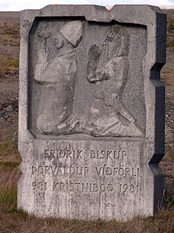A rectangular stone marker with Romanesque-style bas relief depicting two men knelt in prayer. Text below the relief reads: Fridrik Biskup – Þorvaldur víðförli – 981 Kristniboð 1981