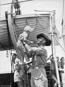 A smiling man in military uniform wearing a slouch hat holds a young child with blonde hair up in the air. Other soldiers are standing in the background under a lifeboat which is suspended on davits above their heads.