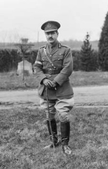 A man in a neat military uniform with a peaked cap, ribbons on his tunic, Sam Brown belt, cane, riding boots and spurs.