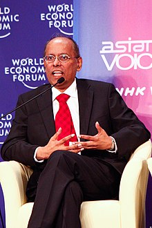 A middle-aged Indian man wearing a black suit, white shirt and red tie speaking into a microphone.