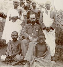 Black and white photo of a seated man wearing a suit, surrounded by several young Ugandan men.