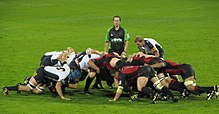 Two opposing formations of eight men, in white and black to the left, red and black to the right, push against each other in a crouched position; behind them stands another player and the referee