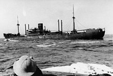 A dark-coloured merchant ship at sea. The head of a person wearing a sun helmet is visible in the foreground