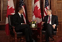 Obama and a man with gray-hair speak in front of American and Canadian flags.