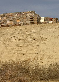 This photo shows a canyon cut into the surrounding flat soil with over 20 distinct horizontal layers of sediment, each clearly demarked from the layer below. Above the canyon lip a number of boxes used for transporting harvested apples are stacked; the size of these boxes indicates that the layers each are 0.5–1 meter in depth.