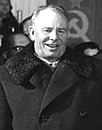 A man wearing a long coat, smiling, in front of a group of people and the Soviet flag