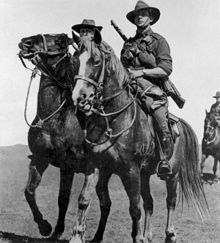 Two men carrying rifles on horseback; another horse and rider are partially visible in the background.