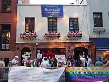 A two-story building with brick on the first floor, with two arched doorways, and gray stucco on the second floor off of which hang numerous rainbow flags.