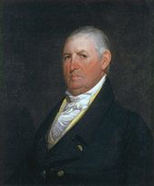 A gray-haired man with a sullen expression wearing a high-collared white shirt, gold vest, and black jacket with gold buttons