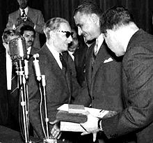 Two men conferring with each other, both are wearing suits and the man on the left is also wearing sunglasses. Three men are standing around them, with one holding a number of objects in his hand