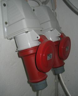 In the foreground, a downward-angled industrial wall socket with a red cap, mounted to a wall; a red plug is inserted into it. In the background, another similar connection.