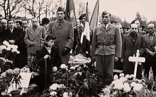Photographie en noir et blanc d'une foule se recueillant dans un cimetière. Au premier plan, une femme accroupie au milieu d'un parterre de bouquets de fleursv, à côté d'une tombe sur laquelle on peut lire « Manoushian ».
