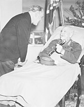A man lies in a hospital bed, wearing an Army uniform instead of pyjamas. His peaked cap is on the blanket and he holds a baton in his hand. A man in a dark suit and pinstripe trousers bends over to talk to him. In the background are flowers, and a flag.