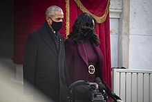 Obama wearing an all-black suit standing next to his wife, both wearing face masks.