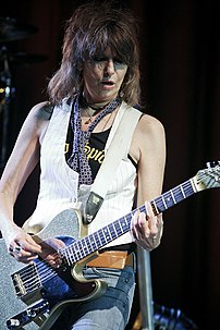 Woman with medium-length shaggy brown hair, wearing a white vest and tie while playing a guitar.