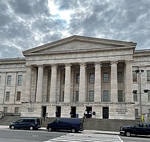 Exterior of a large building made of grey stone. Columns mark the entrance and a wide set of stairs lead up to the columns. Three cars are parked in front of the building. The sun peeks through the cloudy sky above. Several people are walking toward the entrance.