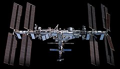 A forward view of the International Space Station with limb of the Earth in the background. In view are the station's sixteen paired maroon-coloured main solar array wings, eight on either side of the station, mounted to a central integrated truss structure. Spaced along the truss are ten white radiators. Mounted to the base of the two rightmost main solar arrays pairs, there are two smaller paired light brown- coloured ISS Roll-out Solar Arrays. Attached to the centre of the truss is a cluster of pressurised modules arranged in an elongated T shape. A set of solar arrays are mounted to the module at the aft end of the cluster.