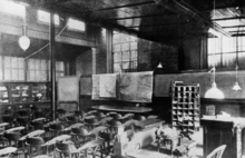 A black and white image of a large room with may chairs in front of a desk and charts. There are bars on the windows.